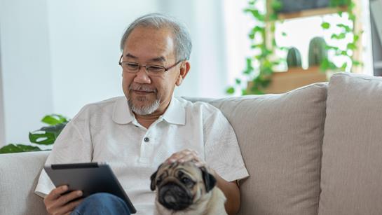 Older Man Sat On The Sofa With His Dog Reading On His Ipad