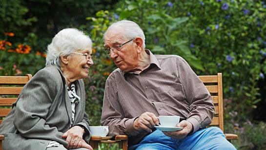 Older Couple Sat On Bench Outside With Cup Of Tea