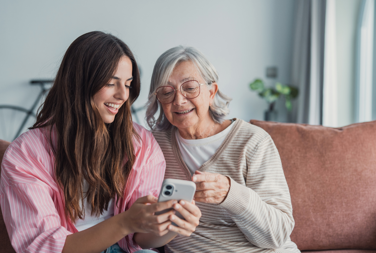 Older Woman At Home Sat On A Sofa Talking To A Younger Woman While Looking At A Mobile Phone