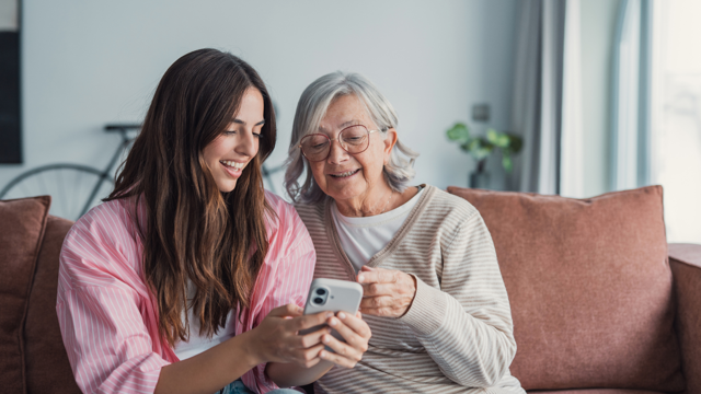 Older Woman At Home Sat On A Sofa Talking To A Younger Woman While Looking At A Mobile Phone
