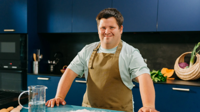 Young Man Stood In The Kitchen Smiling With An Apron On