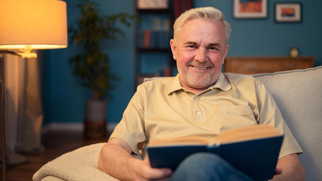 Older Man Reading A Book At Home Sat On The Sofa